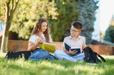 İki genç öğrenci çimlerin üzerinde oturuyor, kitap tutuyor ve dışarıda ders çalışıyor. Öğrenim ve eğitim kavramı