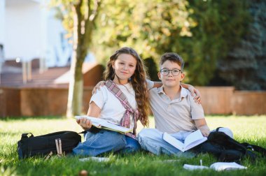 Okul çocukları gülümsüyor, yeşil çimenlerin üzerinde kitap ve sırt çantalarıyla birlikte oturuyor, açık havada öğreniyor ve sosyalleşiyorlar.