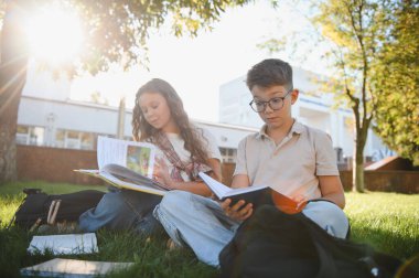 Okul çocukları ders kitapları okuyorlar teneffüste yeşil çimlerin üzerinde oturuyorlar, sırt çantaları güneşte öğreniyor.