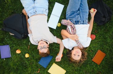 Kitaplar ve elmalarla yeşil çimlerde uzanan iki genç öğrenci, arkadaşlığın ve okuldan uzaklaşmanın keyfini çıkarıyorlar.
