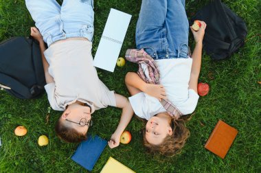 Yeşil çimlerin üzerinde yatan iki okul çocuğu, ders kitapları, sırt çantaları ve elmalarla çevrili, okuduktan sonra dinleniyorlar.