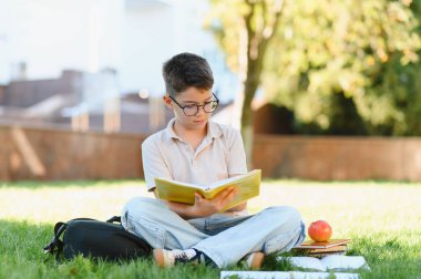 Gözlüklü genç okul çocuğu yeşil kampüs çimlerinde oturmuş kitap okuyor, yanında sırt çantası ve elmayla.
