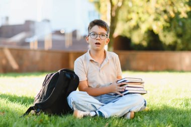 Okul çocuğu elinde kitap ve sırt çantasıyla yeşil çimlerde oturuyor. Kampüste öğrenim görürken şaşırmış görünüyordu.