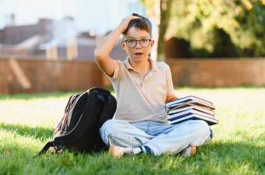 Kitapları ve sırt çantasını tutarken kafası karışmış ve hayal kırıklığına uğramış görünen genç bir öğrenci kampüs çimlerinde oturuyor ve öğreniyor.