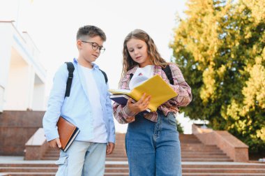 Okul çocukları sırt çantaları ve kitaplar taşıyorlar, dışarıda duruyorlar. Kız açık bir kitap tutuyor, erkek bakıyor. Öğrenim ve eğitim