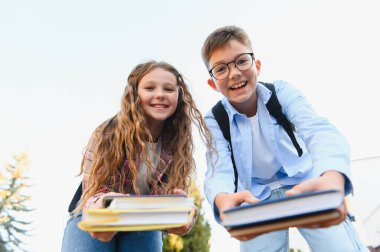 Gülümseyen okul çocuğu ve kız öğrencileri ders kitaplarını tutuyorlar, onları ileri doğru uzatıyorlar, öğrenme ve arkadaşlığı temsil ediyorlar.