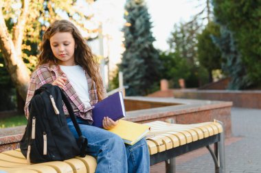 Genç bir kız öğrenci bankta oturuyor, kitap tutuyor ve dışarıda sırt çantasında eşyalar düzenliyor.