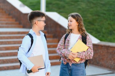İki genç öğrenci, bir erkek ve bir kız, gülümsüyor ve okul kampüsünde tartışıyorlar, ellerinde kitaplar ve sırt çantaları var.