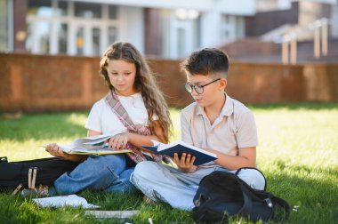Okul çocukları yeşil çimlerin üzerinde oturuyor kitap okuyor, öğreniyor ve bir okul binasının dışında doğal ışık altında iş birliği yapıyorlar.