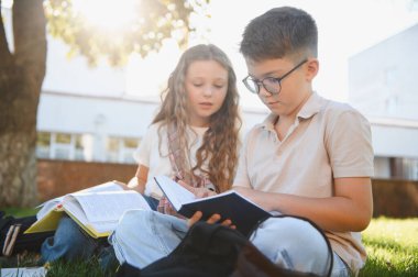 İki okul çocuğu çimlerin üzerinde oturuyor, kitap okuyor ve öğreniyorlar. Açık havada eğitim görmek eğitim için yeni bir ortam sağlar.