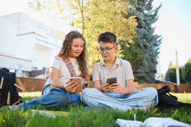 Çimenlerde oturan öğrenciler, ellerinde akıllı telefonlar ve okul molasında açık havada teknolojiyle uğraşıyorlar.