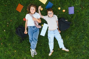 Etrafı kitaplarla, çantalarla ve elmalarla çevrili yeşil çimenlerin üzerinde uzanan iki mutlu öğrenci öğrenmeye ara vermenin keyfini çıkarıyorlar.