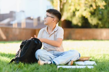 Gözlüklü öğrenci kampüs çimlerinde oturuyor, sırt çantasını açıyor ve çalışmaya hazırlanıyor. Eğitim kavramı