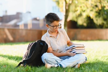 Çimenlerde oturan, elinde ders kitapları ve sırt çantasıyla okula ara verip ders çalışan genç bir öğrenci.