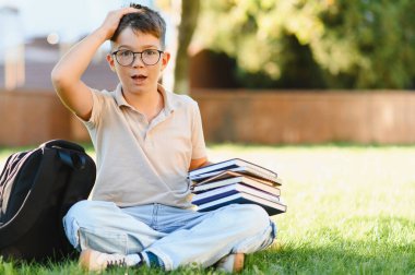 Elinde kitaplarla kampüs çimlerinde oturan gözlüklü bir öğrenci unutulmuş görevler hakkında hayal kırıklığına uğramış bir jest yapıyor.