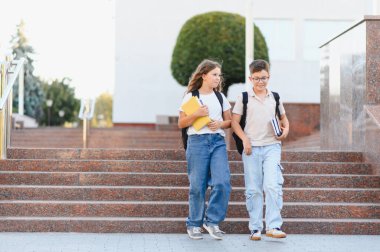 İki okul çocuğu, bir oğlan ve bir kız, kampüsten taş merdivenlerden iniyorlar, kitap taşıyorlar ve sırt çantaları takıyorlar.