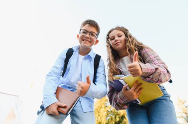 Sırt çantalı gülümseyen öğrenciler kampüste başparmak kaldırıyor, dostluğu, güveni ve akademik başarıyı kutluyorlar.