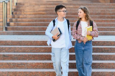 Genç öğrenciler kitap ve sırt çantası taşıyorlar, okul merdivenlerinden iniyorlar ve konuşuyorlar, eğitimi ve arkadaşlığı temsil ediyorlar.