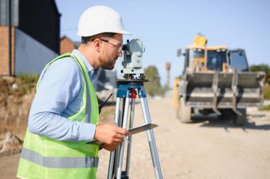 Erkek mühendis, sert şapka ve güvenlik yeleği giyiyor. Açık hava inşaat alanındaki bir araştırma aletine bakıyor.