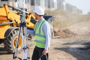 İnşaat alanındaki arazi araştırması için teodolit kullanan sert şapka ve yansıtıcı yelek giyen mühendis.