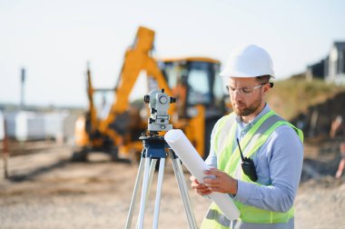 Bir teodolit ve planlarla çalışan genç bir arazi mühendisi, yeni bir inşaat alanı inşaatında hassasiyet ve planlama sağlıyor.