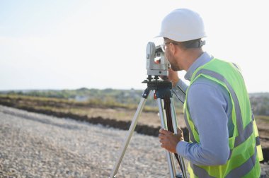 Bir inşaat alanında arazi ölçümü ve haritası için üç ayaklı bir istasyon kullanarak inşaat mühendisi, sert şapka ve yelek giyiyor.