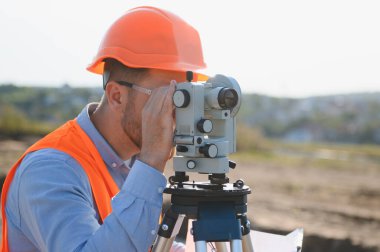 İnşaat mühendisi, sert şapka ve güvenlik yeleği giyiyor, dışarıdaki bir araştırma aracına bakıyor, kesin jeodezik ölçümler yapıyor.