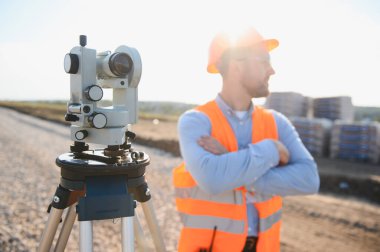 Güvenlik yeleği ve kasket takan profesyonel arazi araştırmacısı. İnşaat alanında teodolit bir aletin yanında kolları bağlı duruyor.