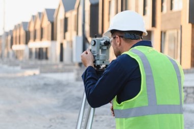 Arazi ölçümü ve yerleşim planlaması için teodolit kullanan sert şapka ve güvenlik yeleği giyen bir araştırmacı.