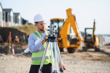 Sert şapka ve güvenlik yeleği giyen genç bir arazi mühendisi inşaat alanının planlarıyla birlikte araştırma ekipmanları yürütüyor.