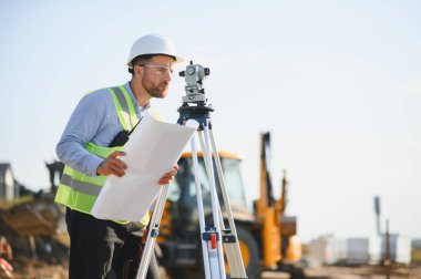 Başlık ve güvenlik yeleği giyen mühendis, inşaat alanındaki arazi araştırmaları için bir teodolit ile planları kontrol ediyor.