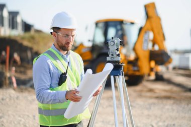 Bir inşaat sahasında çalışan profesyonel bir araştırmacı, arka planda bir teodolit ve kazıcı ile ayrıntılı planları dikkatle inceliyor.