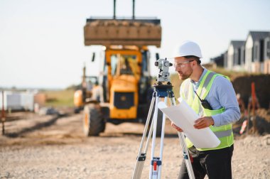 Sert şapka ve güvenlik yeleği giymiş bir adam dışarıda bir teodolitle çalışıyor, elinde planlar tutuyor ve devam eden binayı denetliyor.
