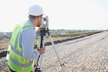 Sert şapka ve güvenlik yeleği giyen arazi mühendisi inşaat alanındaki tüm istasyonu kullanarak yol yapımı için tam ölçüm alanı oluşturdu.