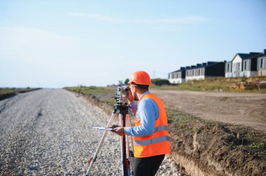 Güvenli yelek giyen profesyonel arazi araştırmacısı ve yeni bir inşaat sahasında arazi ölçümü yapan sert şapkalı.