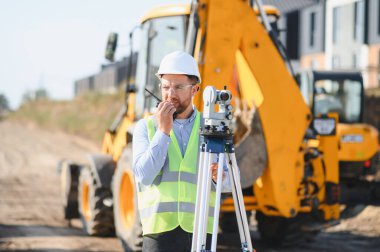 Sert şapka ve güvenlik yeleği giyen erkek arazi mühendisi, tüm istasyon ve ağır inşaat ekipmanlarının yanında telsiz kullanıyor.