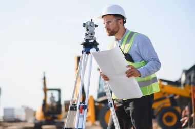 Güvenlik yeleği ve kasket takan mühendis, elinde bir plan var. İnşaat alanındaki toplam bir istasyonu kullanarak araziyi ölçüyor.