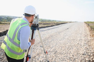 Güvenlik kaskı ve yeleği takan mühendis, altyapı geliştirme çalışmaları için çakıllı bir yolda istasyon kullanıyor.