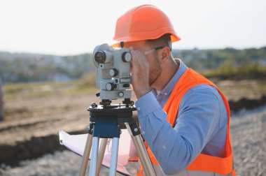 Açık hava şantiyesinde güvenlik yeleği ve şapka takan mühendis bir inceleme aleti kullanıyor.