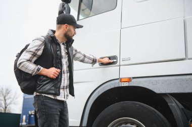 Şapka ve sırt çantası takmış genç bir adam büyük beyaz bir tırın kapısını açıyor ve günlük yolculuğuna hazırlanıyor.