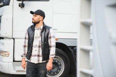 Genç bir kamyon şoförü büyük beyaz bir kamyonun yanında duruyor. Kameraya bakmıyor, kep ve yelek giyiyor.