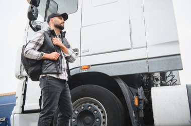 Kamyon şoförü sırt çantası taşıyor, modern ticari bir tırın yanında duruyor, ileriye bakıyor, uzun bir yolculuğa hazır.