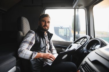 Yarı kamyon kabininde oturan, dizüstü bilgisayarda yazan, lojistik ve ulaşım işlemlerini yöneten profesyonel kamyon şoförü.