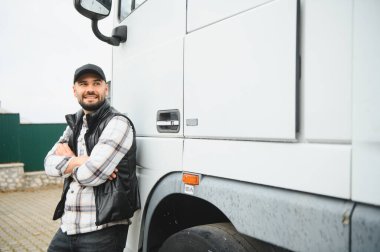 Yelek ve şapka giyen genç bir kamyon şoförü beyaz kamyonetinin yanında çapraz kollarıyla güvenle duruyor.