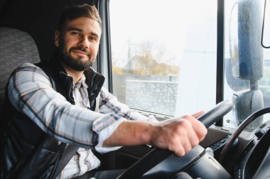 Sakallı ve ekose gömlekli bir adam bir kamyon kabininde emniyet kemeri takıyor, direksiyonu tutuyor, kameraya bakıyor.