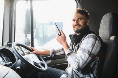 Kamyon şoförü aracın içinde bir iletişim cihazı tutuyor, gülümsüyor ve dışarı bakıyor, lojistik ve ulaşımı temsil ediyor.