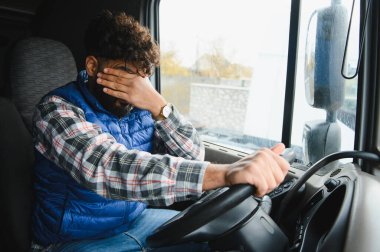 Kamyon şoförü, kabinin içinde kamyon sürerken yorgun, stresli ve baş ağrısı olduğunu belirterek yüzünü kapatıyor.