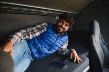 Çelik yelek ve ekose gömlek giyen mutlu genç bir adam bir karavan yolculuğunda bir karavanda dinleniyor.