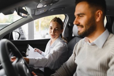 Araba satıcısı kadın, yeni bir arabada oturan erkek müşteriye özelliklerini anlatıyor. Satış hizmeti ve araç alımı