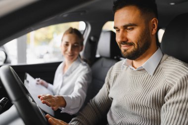 Bir kadın satıcı otomobil galerisinde bilgi sağlarken, erkek arabanın iç detaylarını inceliyor